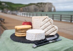 Four famous cheeses of Normandy, squared pont l'eveque, camembert cheese, yellow livarot, heartshaped neufchatel and view on promenade and alebaster cliffs Porte d'Aval in Etretat, Normandy, France
